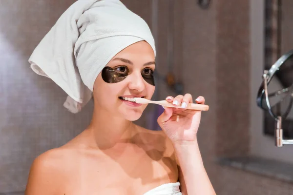 woman dressed in a towel to brush her teeth in front of a mirror in the bathroom. healty wellness morning concept