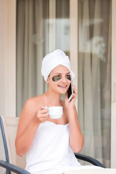 Young woman under-eye patches talking by phone and drinking coffee at hotel terrace resort