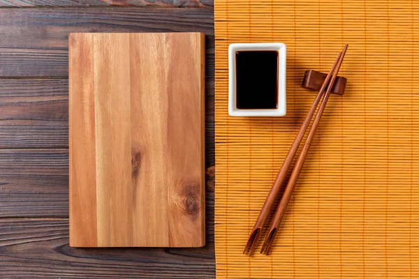 Placa de madera rectangular vacía con palillos para sushi y salsa de soja sobre fondo de madera. Vista superior con espacio de copia —  Fotos de Stock