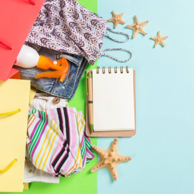 Renkli alışveriş çantaları giysi dolu yaz konsepti. T-shirt, kot şort, dizüstü bilgisayar ve güneş kremi şişesi ile hediye çantaları. Yaz dilek listesi kavramı. Mavi arka planda üst görünüm kopyalama alanı