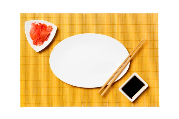 Placa blanca ovalada vacía con palillos para sushi y salsa de soja, jengibre sobre fondo de estera de bambú amarillo. Vista superior con espacio de copia para su diseño —  Fotos de Stock