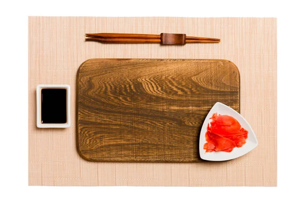 Placa de madera marrón rectangular vacía con palillos para sushi, jengibre y salsa de soja sobre fondo de estera de sushi marrón. Vista superior con espacio de copia para su diseño —  Fotos de Stock