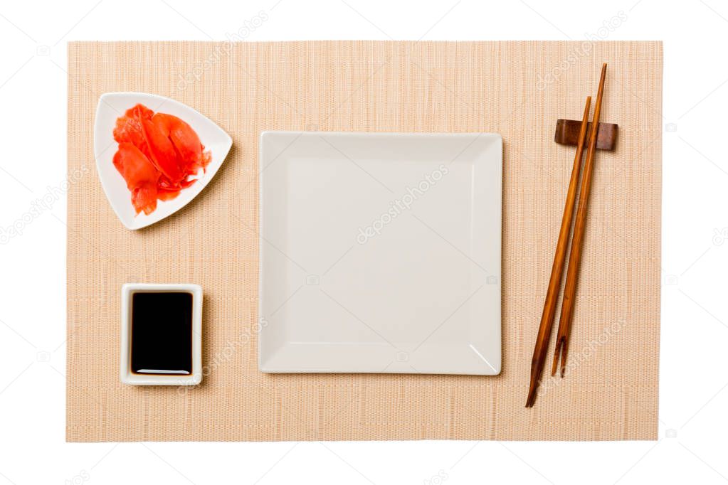 Empty white square plate with chopsticks for sushi and soy sauce, ginger on brown sushi mat background. Top view with copy space for you design