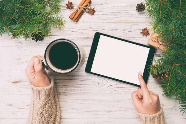 Christmas online shopping. Female hand touch screen of tablet, top view on wooden bakground, copy space and holding mug of coffee. Winter holidays sales background. Toned