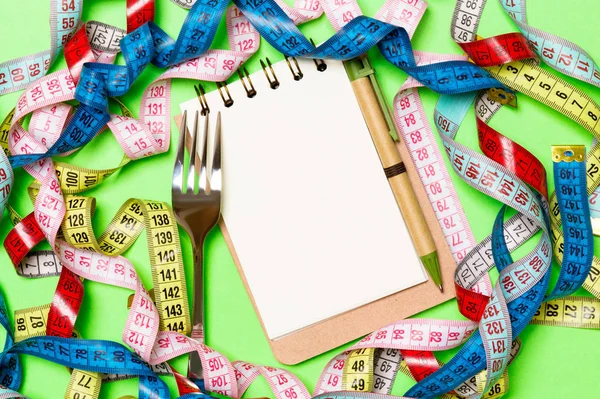 Gruppo di nastri di misura colorati, penna, notebook aperto e forchetta su sfondo verde con spazio vuoto per la tua idea. Vista dall'alto del concetto di stile di vita sano — Foto Stock