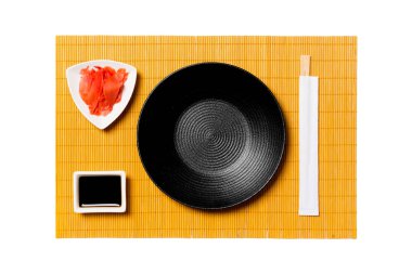 Suşi ve soya sosu için çubukları ile Boş yuvarlak siyah plaka, sarı bambu mat arka plan üzerinde zencefil. Tasarım için kopyalama alanı ile üst görünüm