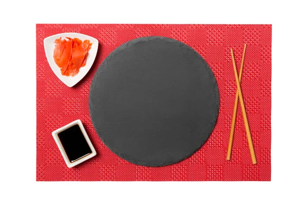 Emptyround zwarte leisteen plaat met eetstokjes voor sushi en sojasaus, gember op rode mat Sushi achtergrond. Bovenaanzicht met Kopieer ruimte voor uw ontwerp — Stockfoto