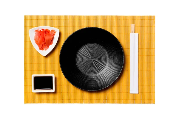 Tom rund svart tallrik med ätpinnar för sushi och sojasås, ingefära på gul bambu matta bakgrund. Toppvy med kopierings utrymme för dig design — Stockfoto