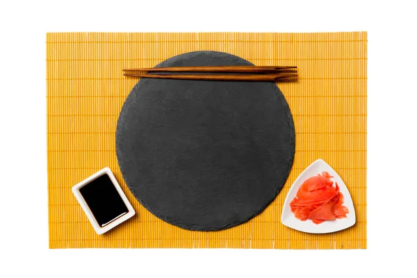 Leerer schwarzer Schieferteller mit Essstäbchen für Sushi, Ingwer und Sojasauce auf gelbem Bambusmattenhintergrund. Ansicht von oben mit Kopierraum für Ihr Design — Stockfoto