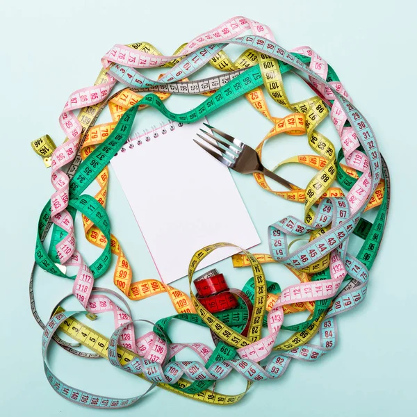 Mavi arka plan üzerinde renkli ölçüm bantları, çatal ve not defteri kompozisyonu. Tasarım için diyet kontrolü ve boş alan üst görünümü