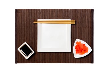 Koyu bambu paspas arka plan üzerinde suşi, zencefil ve soya sosu için çubukları ile Boş beyaz kare plaka. Tasarım için kopyalama alanı ile üst görünüm
