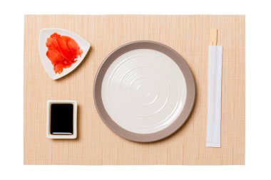 Kahverengi suşi mat arka plan üzerinde suşi, zencefil ve soya sosu için çubukları ile Boş yuvarlak beyaz plaka. Tasarım için kopyalama alanı ile üst görünüm