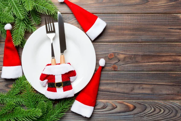 Holiday composition of plate and flatware decorated with Santa hat and clothes on wooden background. Top view of Christmas decorations with empty space for your design. Festive time concept — Stock Photo, Image