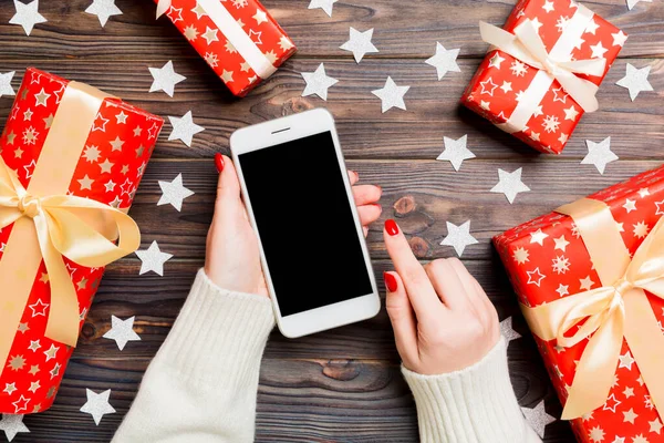 Top view egy nő kezében egy telefont a fa újévi háttér készült ajándékdobozok és hópelyhek. Karácsonyi ünnepi koncepció. Mockup — Stock Fotó