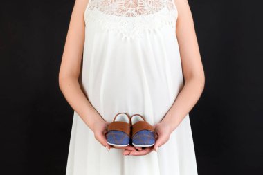 Hamile bir kadının ellerinde siyah arka planda karnına dayanan küçük bir çocuk için kesilmiş küçük ayakkabılar. Gelecekteki çocuğa sevgiler. Boşluğu kopyala.