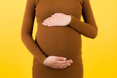 Sarı arka planda hamile karnını kapat. Annem kahverengi bir elbise giyiyor ve karnını tutuyor. Ebeveynlik kavramı. Boşluğu kopyala.