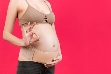 Destekleyici bandaj takan hamile kadına yakın durun ve pembe arka planda fotokopi aletiyle iyi niyet göstergesi gösterin. Ortopedik abdominal destek kemeri kavramı.