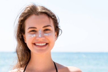 Güzel genç bir kadın kameraya poz veriyor. Yüzünde güneş kremiyle, denizin arka planında..