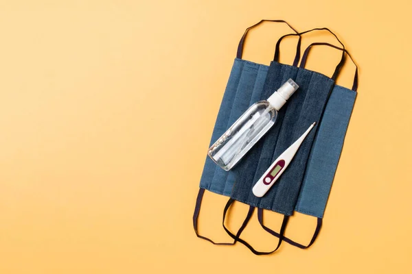 Top view of handmade cotton masks, alcohol hand sanitizer and digital thermometer on orange background with copy space. Protective medical equipment concept.