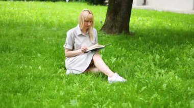 Güneşli bir günde çimlerin üzerinde oturmuş kitap okuyan bir kadın.