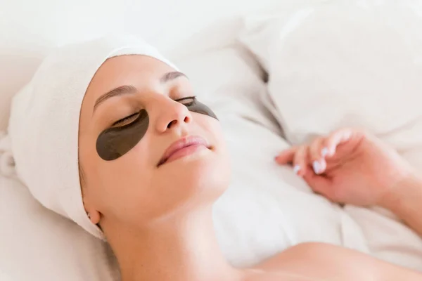 Young woman with closed eyes and black eye patches is lying and relaxing in the bed after having a bath wrapped in towel. Beauty treatment and skincare concept.