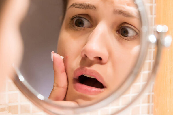 Young girl in front of a bathroom mirror putting cream on a red pimple. Beauty skincare and wellness morning concept.