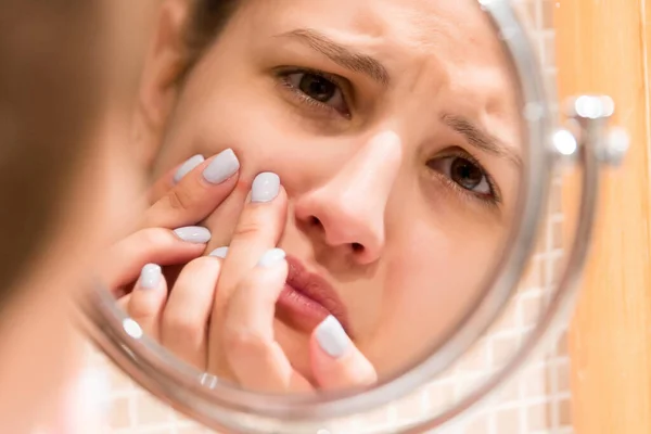 Young girl squeezes pimple on the fer face in front of a bathroom mirror. Beauty skincare and wellness morning concept.