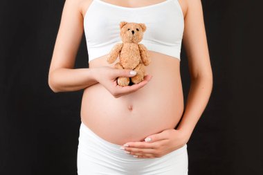 Beyaz iç çamaşırlı hamile kadının siyah arka planda karnına oyuncak ayı dayadığı yakın plan. Genç anne bir bebek bekliyor. Boşluğu kopyala.
