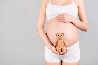 Beyaz iç çamaşırlı mutlu hamile bir kadının gri arka planda karnına oyuncak ayı dayadığı görüntüler. Çocuk bekliyor. Boşluğu kopyala.