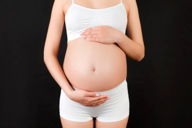 Beyaz iç çamaşırı giyen hamile bir kadın resmi. Genç anne, siyah arka planda bir bebek beklerken karnına sarılıyor. Boşluğu kopyala.