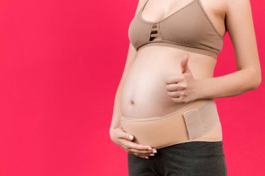 Hamile bir kadının baş parmağını göstermesi ve elastik hamilelik bandajı takıp pembe arka plandaki ağrının kopyalanması için fotokopi çekmesi. Ortopedik abdominal destek kemeri kavramı.