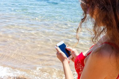 Kırmızı mayo giymiş genç bir kız tatilde cep telefonuyla denizin fotoğrafını çekiyor. Plaj konsepti.