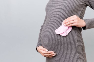 Hamile bir kadının ellerinde gri arka planda göbeğine uzanan pembe çorapların görüntüsü. Mutlu hamilelikler. Boşluğu kopyala.