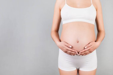 Gri arka planda karnına dokunan hamile kadına yakın çekim. Müstakbel anne beyaz iç çamaşırı giyiyor. Bir bebek bekliyorduk. Boşluğu kopyala.