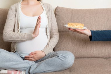 Kanepede oturan hamile kadına yakın çekim. Lezzetli kek dilimlerini reddediyor ve jesti durduruyor. Hamilelik konsepti sırasında diyet.