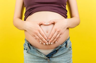 Fermuarsız kot pantolonlu hamile bir kadının renkli bir arka planda ellerini karnının üstünde tutarak fotokopi çekmesine yakın çekim. Annelik kavramı.