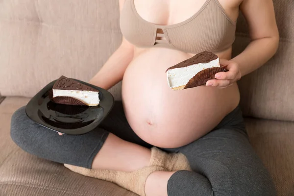 お腹を空かせた妊婦のクローズアップは自宅でソファでリラックスしたおいしいケーキを食べています 妊娠中の甘い渇望 — ストック写真