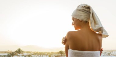 Duş aldıktan sonra beyaz havluya sarılı bir şekilde balkonda duran genç bir kadının arka görüntüsü. Kız dağ manzarası ve gün batımının tadını çıkarıyor. Temiz ve taze konsept.