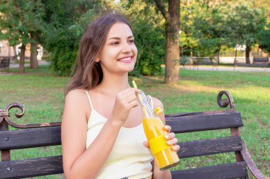 Genç güzel kız parkta bankta dinleniyor ve pipetten taze meyve suyu içiyor..
