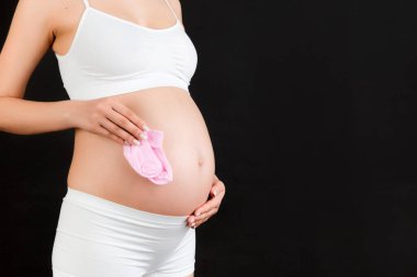 Siyah arka planda bir bebek için pembe çorap tutan beyaz iç çamaşırlı hamile kadın portresi. Çocuk doğurmak üzere. Boşluğu kopyala.