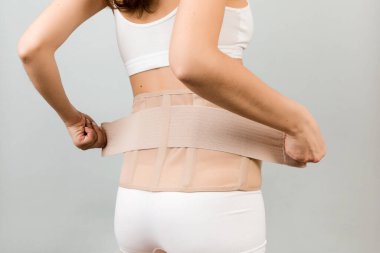 Back view of pregnant woman in underwear putting on supporting bandage to reduce backache at gray background with copy space. Cropped image of orthopedic abdominal support belt concept. clipart