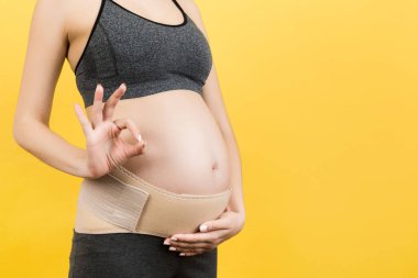 Hamile bir kadının sırt ağrısına karşı ortopedik bandajı destekleyin. Sarı arka planda fotokopi alanı olan iyi işareti gösterin. Ortopedik abdominal destek kemeri kavramı.
