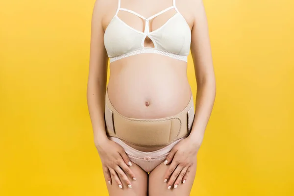 Close up of pregnant woman in underwear with supporting bandage against backache at yellow background with copy space. Orthopedic abdominal support belt concept.