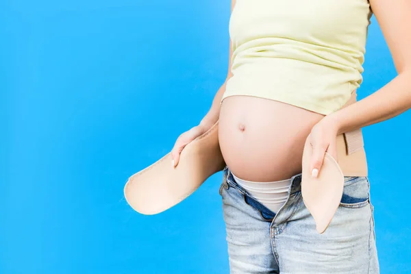 Close up of pregnant woman dressing maternity belt at blue background with copy space. Support belt against backache. Orthopedic abdominal support belt concept.