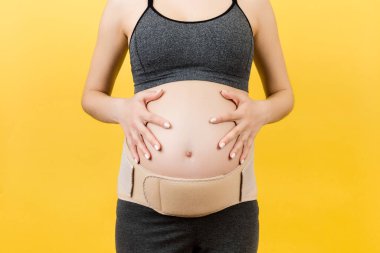 Sarı arka plandaki sırt ağrısını fotokopi aletiyle azaltmak için hamile kadının üzerine giydirilmiş elastik annelik bandını kapatın. Ortopedik abdominal destek kemeri kavramı.