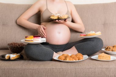 Kanepede oturan aç hamile kadına yakın çekim yapmak, çörek, kek, çikolata gevreği, kruvasan ve kurabiye gibi sağlıksız yiyecekler yemek demektir. Hamilelik konseptinde tatlı aşermeler.