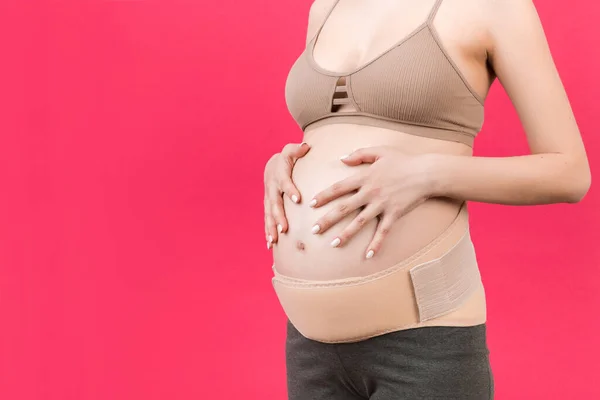 Close up of pregnancy bandage dressed on pregnant woman for reducing backpain at pink background with copy space. Orthopedic abdominal support belt concept.