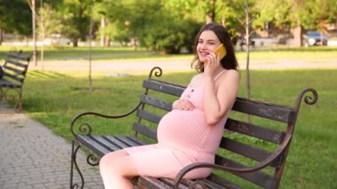 Gülümseyen hamile kadın yaz kenti parkında bankta oturuyor ve cep telefonuyla konuşuyor.