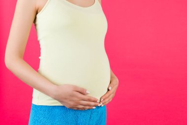 Karnına sarılan hamile bir kadın görüntüsü. Annem pembe arka planda renkli ev kıyafetleri giyiyor. Müstakbel ebeveyn çocuğunu bekliyor. Boşluğu kopyala.