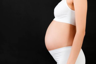 Siyah arka planda hamile bir kadının göbeğinin yakın portresi. Annem beyaz iç çamaşırı giyiyor. Annelik kavramı. Boşluğu kopyala.